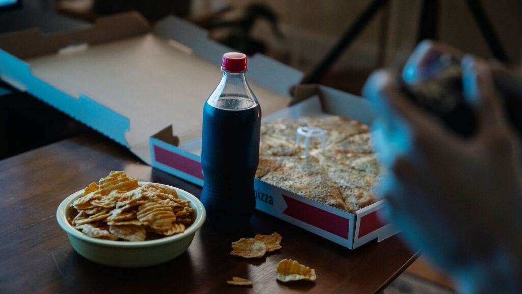 Crop unrecognizable person at desk with bottle of soft drink between crunchy potato chips and takeaway pizza at home