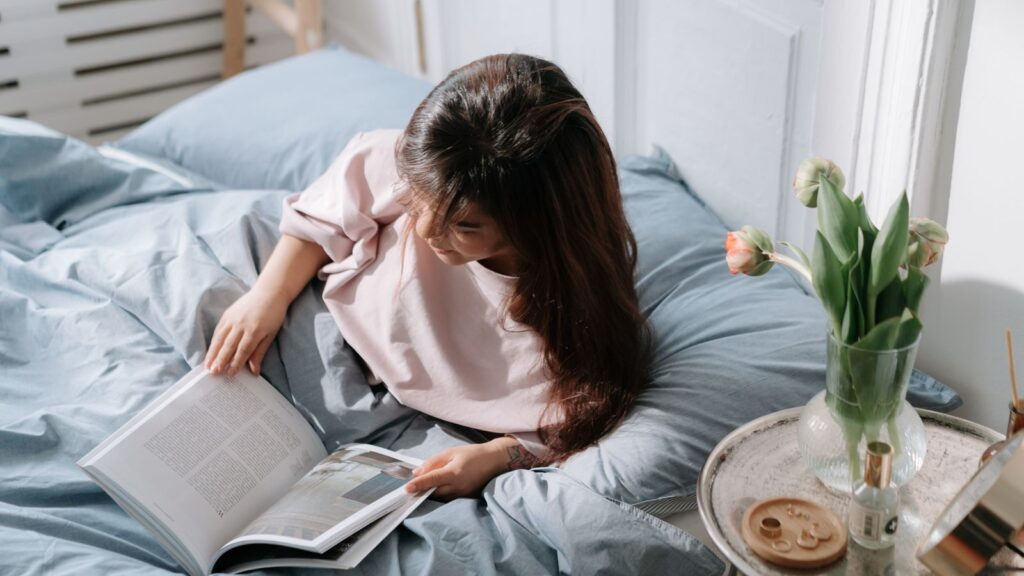 An Asian woman enjoying a peaceful morning reading in a cozy bedroom with soft light and floral decor.