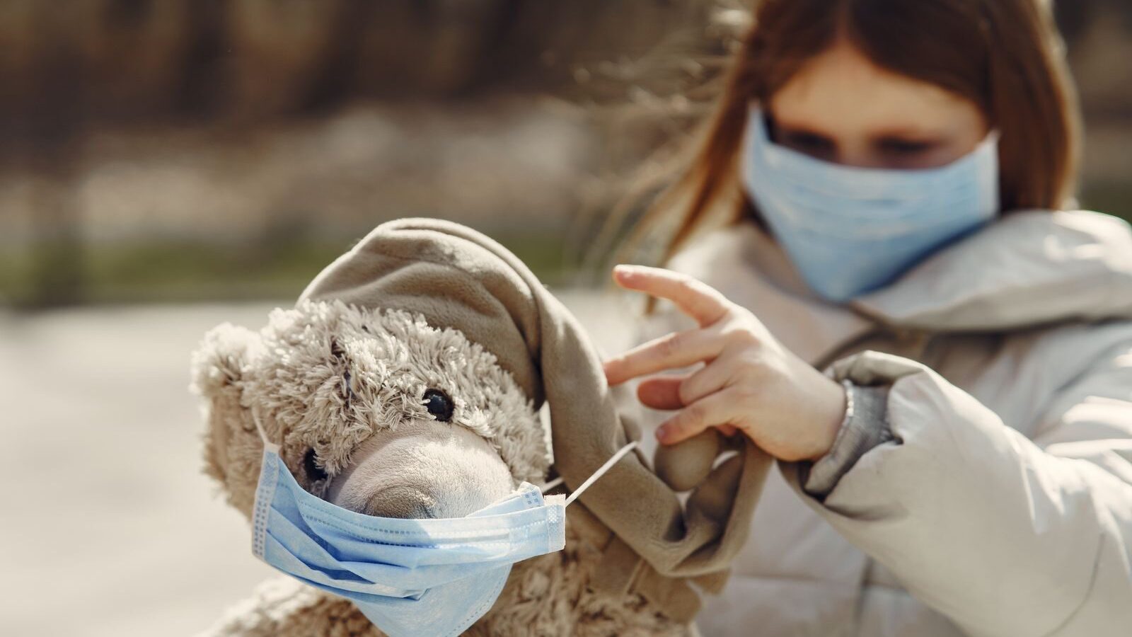 Child in warm outerwear and protective mask putting medical mask on favorite plush toy while spending time in autumn park