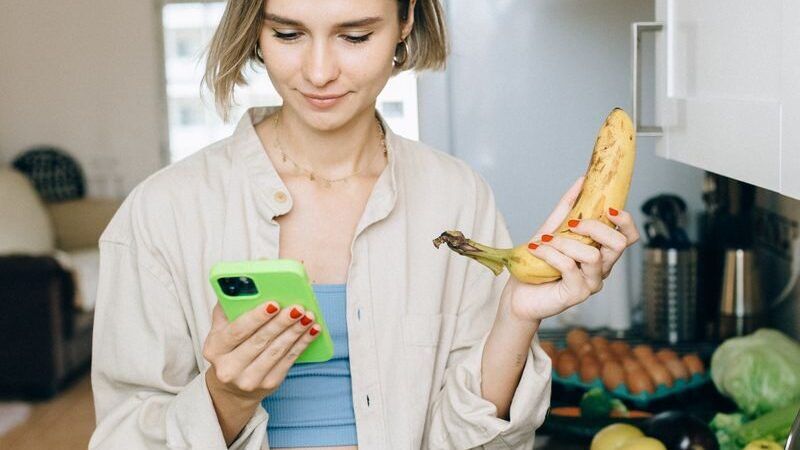 Young woman checks her smartphone while holding a banana, standing by groceries in the kitchen.