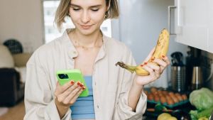 Young woman checks her smartphone while holding a banana, standing by groceries in the kitchen.